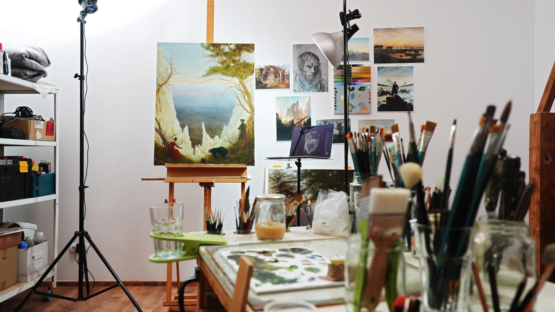 Auf einer Staffelei in einem Atelier ist ein Gemälde aufgestellt, auf dem eine Landschaft mit einer weißen Felsenbucht erkennbar ist.