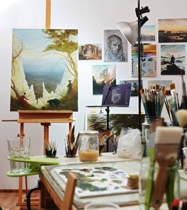Auf einer Staffelei in einem Atelier ist ein Gemälde aufgestellt, auf dem eine Landschaft mit einer weißen Felsenbucht erkennbar ist.
