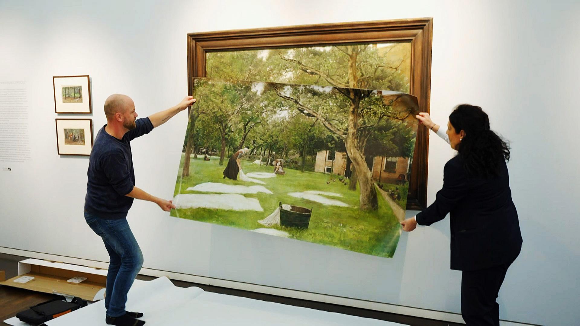 Ein Mann hält mit einer Frau ein großes Plakat vor einem aufgehängten Bild. Darauf sind Bäume und ein Haus zu sehen, sowie zwei Wäscherinnen die ihre weiße Wäsche auf den Rasen auslegen.