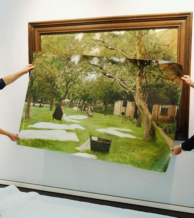 Ein Mann hält mit einer Frau ein großes Plakat vor einem aufgehängten Bild. Darauf sind Bäume und ein Haus zu sehen, sowie zwei Wäscherinnen die ihre weiße Wäsche auf den Rasen auslegen.