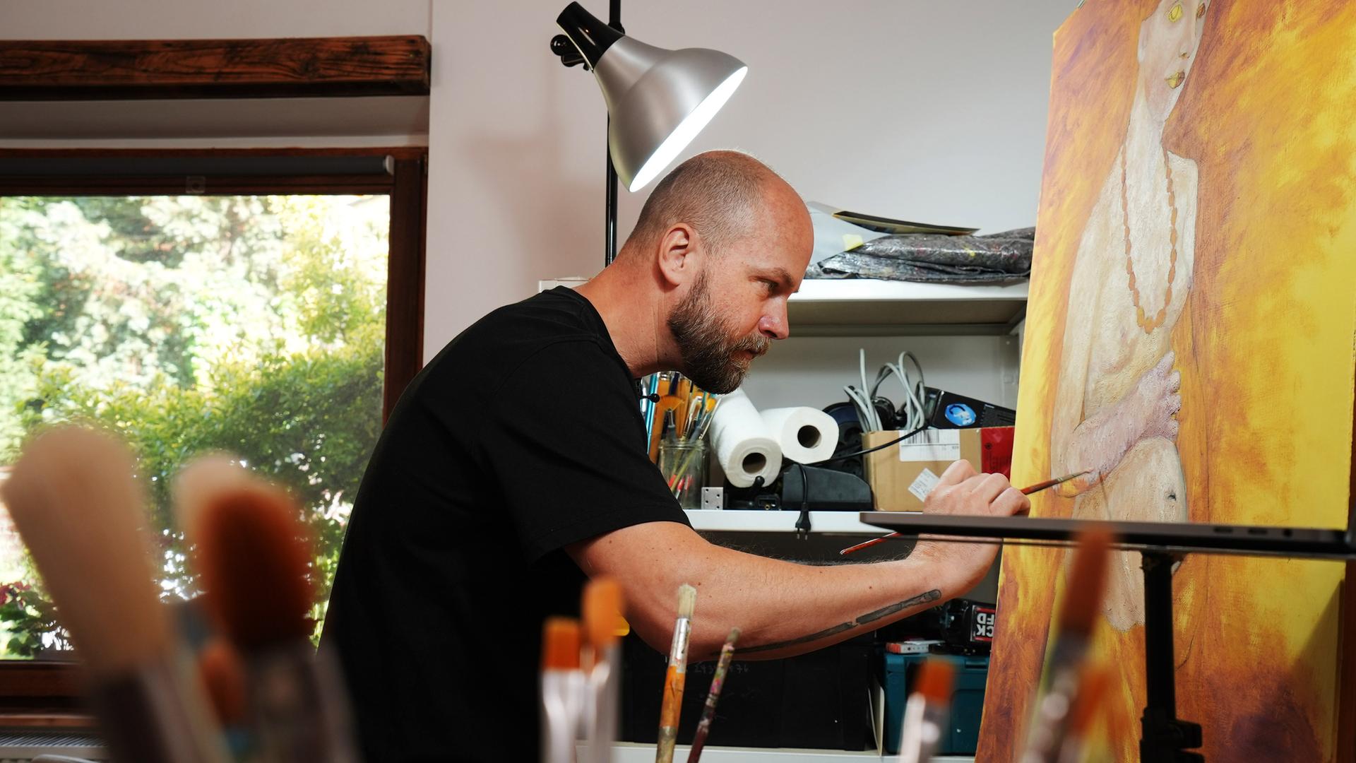 Ein Mann in schwarzem T-Shirt ist im Profil fotografiert, während er auf eine Leinwand malt. Darauf ist ein nackter, schwangerer Oberkörper erkennbar, vor einem einen gold-gelblichen Hintergrund.