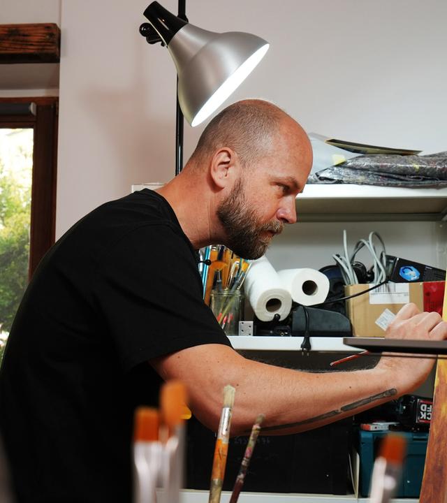 Ein Mann in schwarzem T-Shirt ist im Profil fotografiert, während er auf eine Leinwand malt. Darauf ist ein nackter, schwangerer Oberkörper erkennbar, vor einem einen gold-gelblichen Hintergrund.