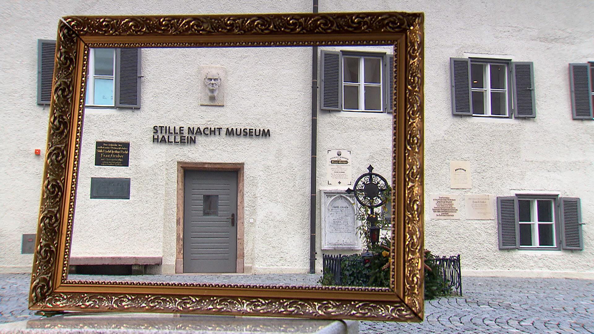 Das Bild zeigt den Eingang zum „Stille Nacht Museum“ in Hallein, Österreich. Der Eingang ist mit einer grauen Tür gestaltet, über der ein Schild mit der Aufschrift „STILLE NACHT MUSEUM HALLEIN“ angebracht ist. An der Wand über der Tür befindet sich ein reliefartiges Gesicht, das vermutlich eine historische Figur darstellt.   Rechts neben dem Eingang sind mehrere Schilder und Plaketten angebracht, die Informationen über das Museum enthalten. Der Bereich vor dem Eingang ist gepflastert und mit einem kleinen, verzierten Gitterzaun sowie Pflanzen dekoriert.   Im Vordergrund des Bildes ist ein goldener Bilderrahmen zu sehen, der einen Teil der Fassade des Museums umrahmt. Die Gebäude sind typisch für die Region, mit weißen Wänden und Fenstern, die mit grünen Fensterläden versehen sind.