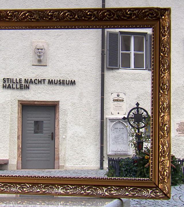 Das Bild zeigt den Eingang zum „Stille Nacht Museum“ in Hallein, Österreich. Der Eingang ist mit einer grauen Tür gestaltet, über der ein Schild mit der Aufschrift „STILLE NACHT MUSEUM HALLEIN“ angebracht ist. An der Wand über der Tür befindet sich ein reliefartiges Gesicht, das vermutlich eine historische Figur darstellt.   Rechts neben dem Eingang sind mehrere Schilder und Plaketten angebracht, die Informationen über das Museum enthalten. Der Bereich vor dem Eingang ist gepflastert und mit einem kleinen, verzierten Gitterzaun sowie Pflanzen dekoriert.   Im Vordergrund des Bildes ist ein goldener Bilderrahmen zu sehen, der einen Teil der Fassade des Museums umrahmt. Die Gebäude sind typisch für die Region, mit weißen Wänden und Fenstern, die mit grünen Fensterläden versehen sind.