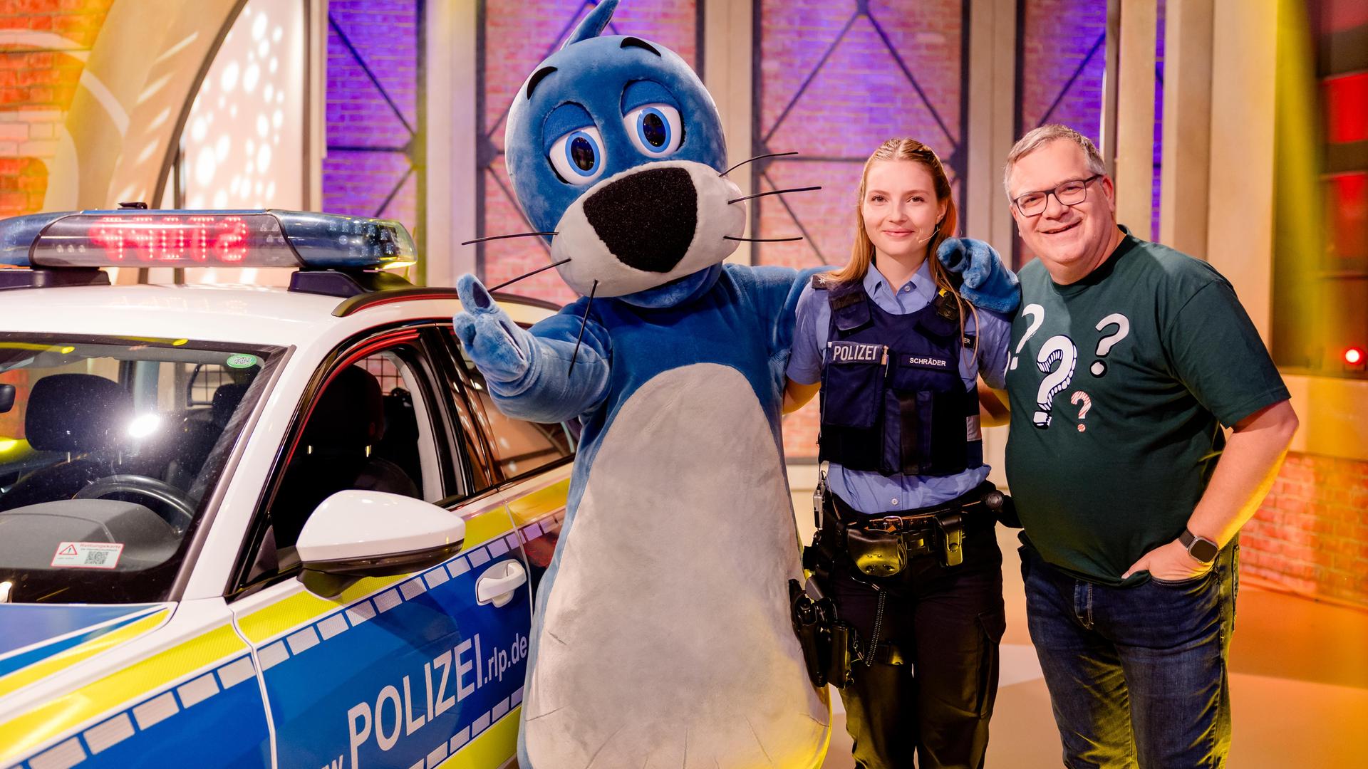 Polizeikommissarin Sarah Schräder steht zwischen Piet Flosse und Elton im Studio. Hinter ihnen steht ein Polizeiauto.