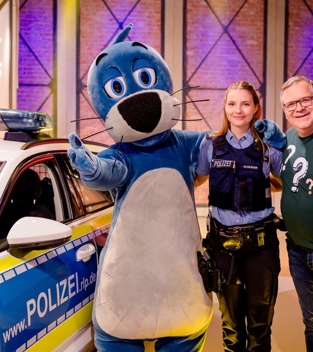Polizeikommissarin Sarah Schräder steht zwischen Piet Flosse und Elton im Studio. Hinter ihnen steht ein Polizeiauto.