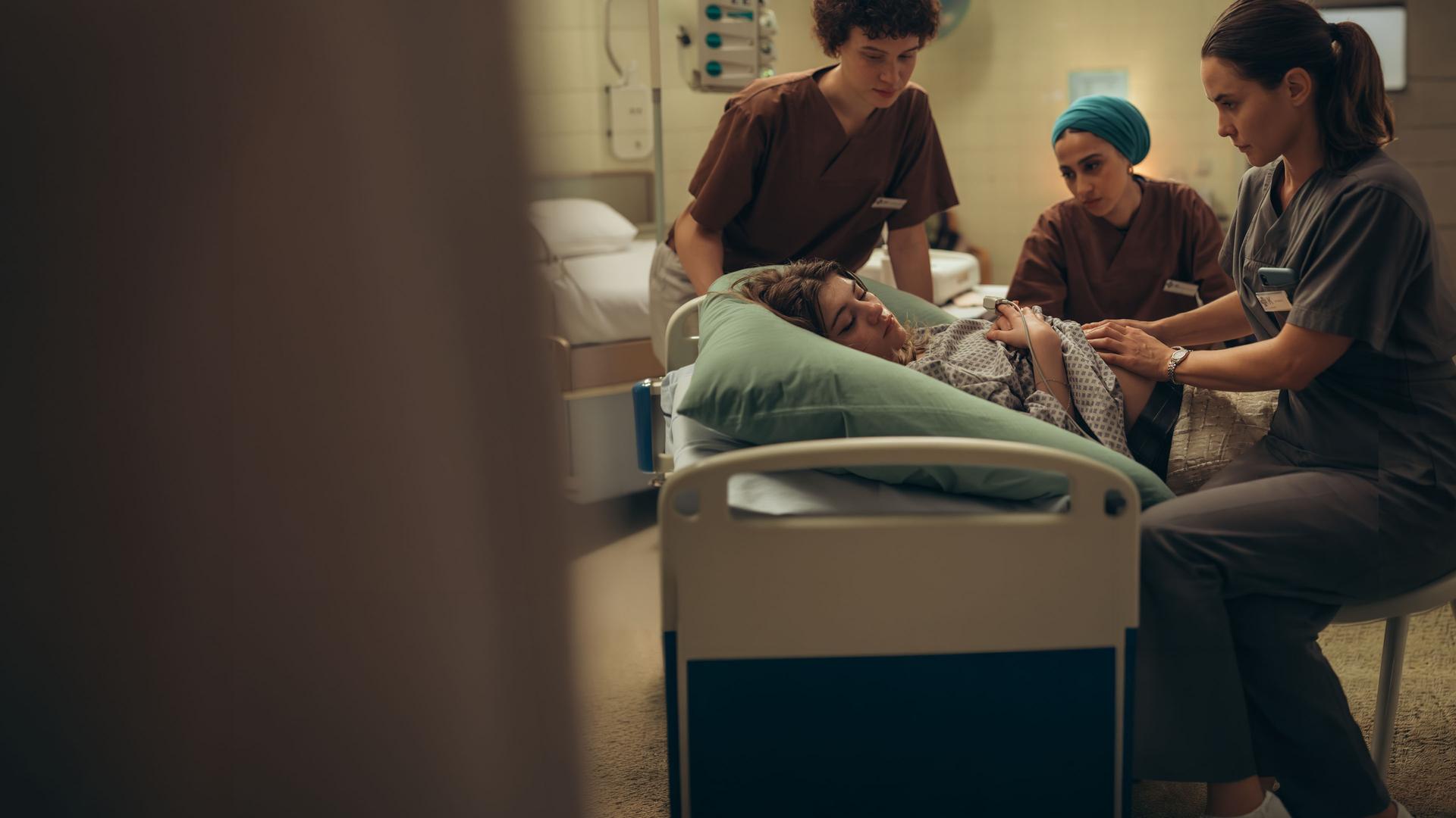 "PUSH - Geheimnisse": Lou (Lisa Moell) liegt in der Klinik. Greta (Lydia Amasko) und Meryem (Tua El-Fawwal) stehen daneben während Dr. Mohn (Katia Fellin) Lou untersucht.