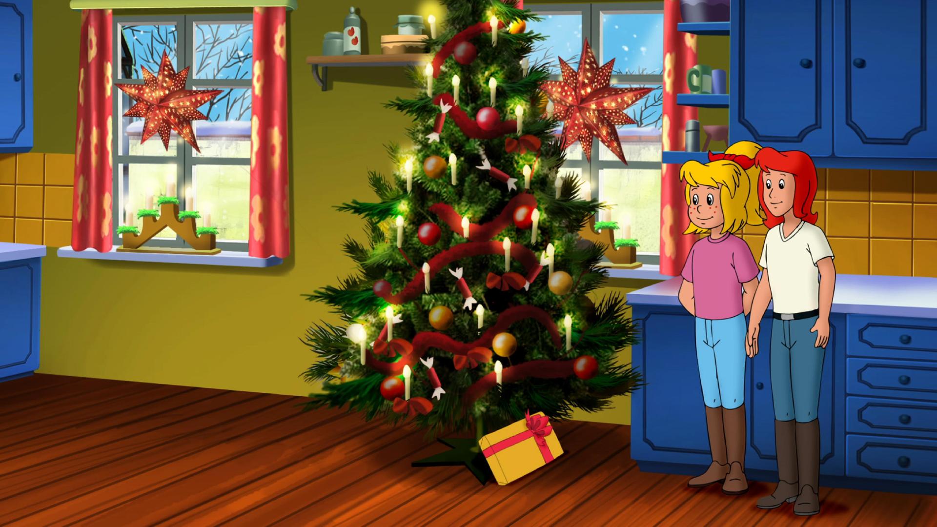 Bibi und Tina (rechts) stehen im Wohnzimmer neben einem geschmückten Weihnachtsbaum (Mitte). Unter dem Baum liegt ein Geschenk mit gelbem Papier und roter Schleife.