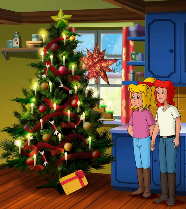 Bibi und Tina (rechts) stehen im Wohnzimmer neben einem geschmückten Weihnachtsbaum (Mitte). Unter dem Baum liegt ein Geschenk mit gelbem Papier und roter Schleife.