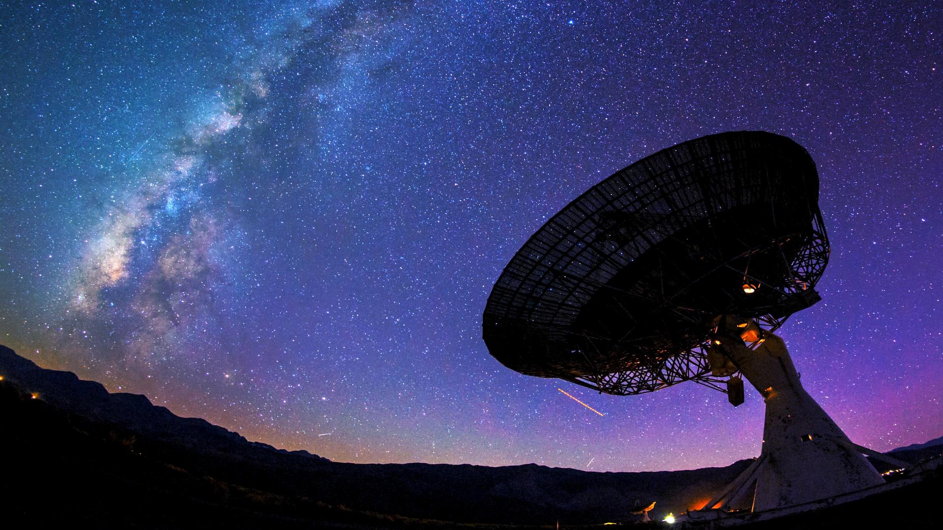 Ein riesiges Teleskop steht vor dem Nachthimmel. Dahinter ist die Milchstraße zu sehen.