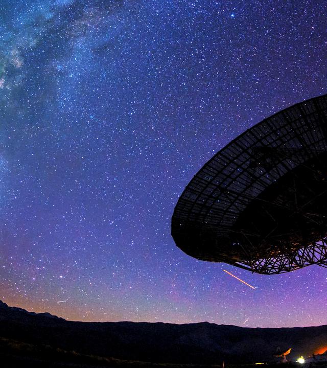 Ein riesiges Teleskop steht vor dem Nachthimmel. Dahinter ist die Milchstraße zu sehen.
