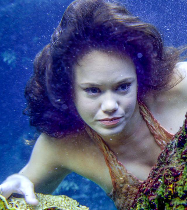 Meerjungfrau Mimmi versteckt sich unter Wasser hinter einem Felsen und beobachtet etwas.