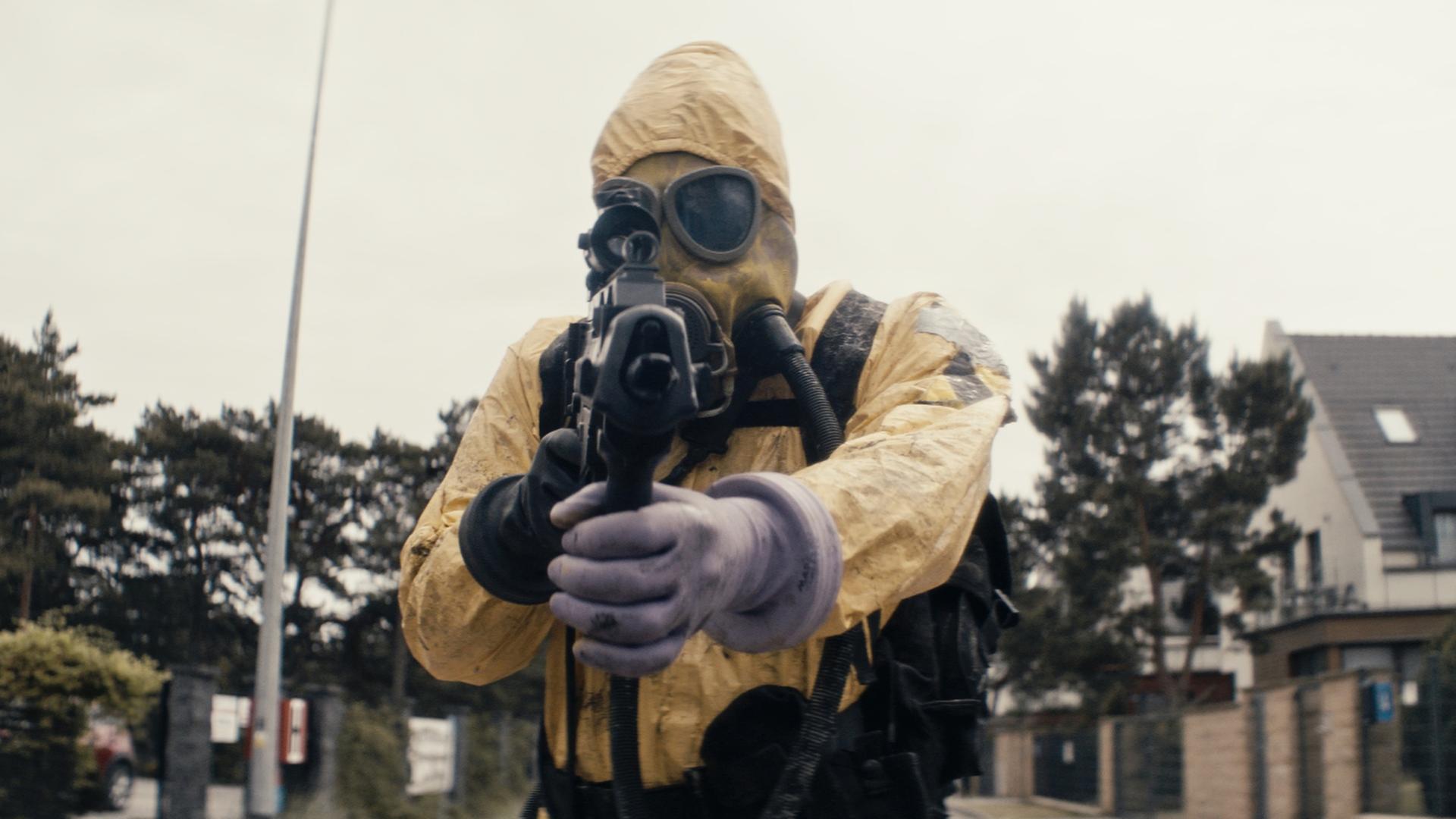 Ein Mann in einem gelben Ganzkörperanzug und mit einer Atemmaske auf dem Gesicht steht auf einer Straße und richtet seine Waffe auf die Kamera.