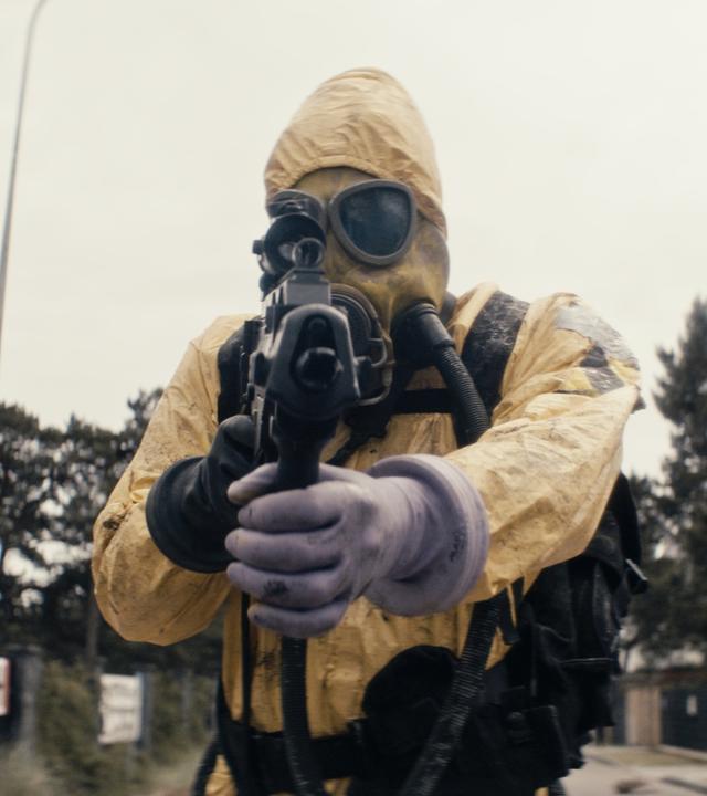Ein Mann in einem gelben Ganzkörperanzug und mit einer Atemmaske auf dem Gesicht steht auf einer Straße und richtet seine Waffe auf die Kamera.
