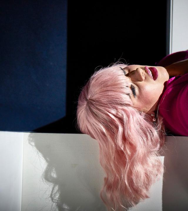 Das Bild zeigt eine weibliche Person mit langen, welligen, platinblonden Haaren, die in einer entspannten Position auf einer weißen Fläche liegt. Sie trägt einen auffälligen, pinken Mantel und darunter ein Kleid mit Leopardenmuster. Ihr Gesicht ist leicht zur Seite geneigt, und ihre Lippen sind leicht geöffnet, was einen nachdenklichen oder melancholischen Ausdruck vermittelt.  Neben ihr steht ein Glas mit einem Drink, in dem sich eine Zitronenscheibe befindet. Der Hintergrund ist dunkel, was einen interessanten Kontrast zur hellen Kleidung der Person bietet. Der Lichteinfall schafft Schatten, insbesondere um das Gesicht und die Haare der abgebildeten Person, und verstärkt den dramatischen Eindruck der Szene.