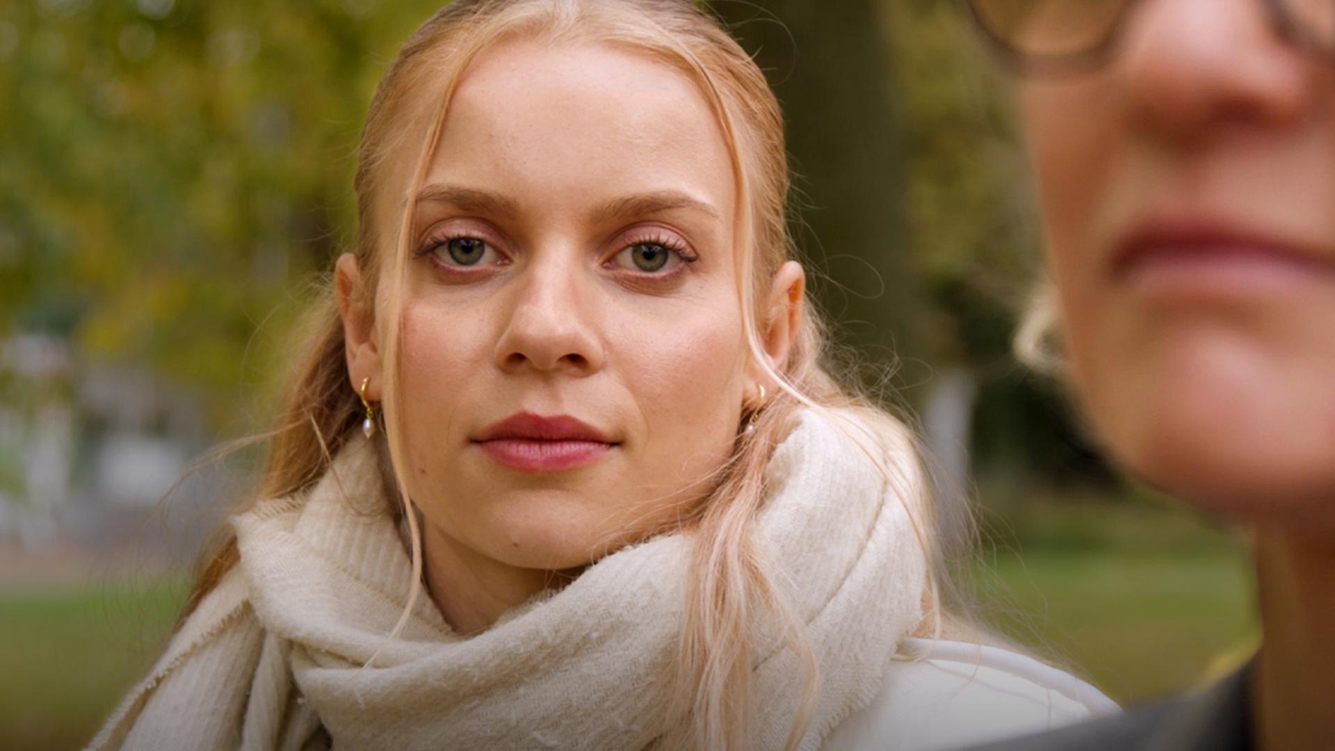 Im Zentrum des Bildes eine junge Frau mit blonden Haaren, die in die Kamera blickt
