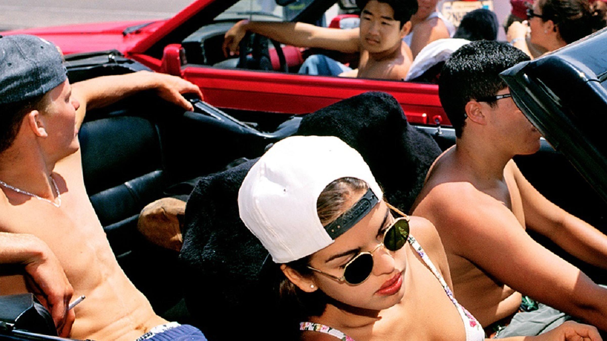 Cabriolet mit gelangweilten Jugendlichen. Auf dem Fahrer- und dem Rücksitz sitzen Jungs. Auf dem Beifahrersitz ein schönes Mädchen im Bikinioberteil,.