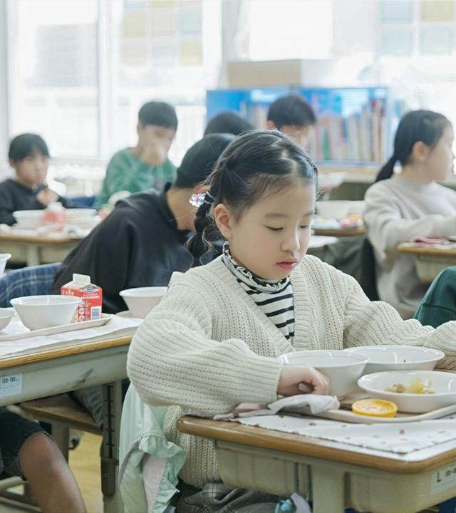Schülerinnen und Schüler essen konzentriert ihr Mittagessen an ihren Schreibtischen in einem hellen Klassenzimmer. Jeder hat ein Tablett mit mehreren Schalen und einem Getränk vor sich.
