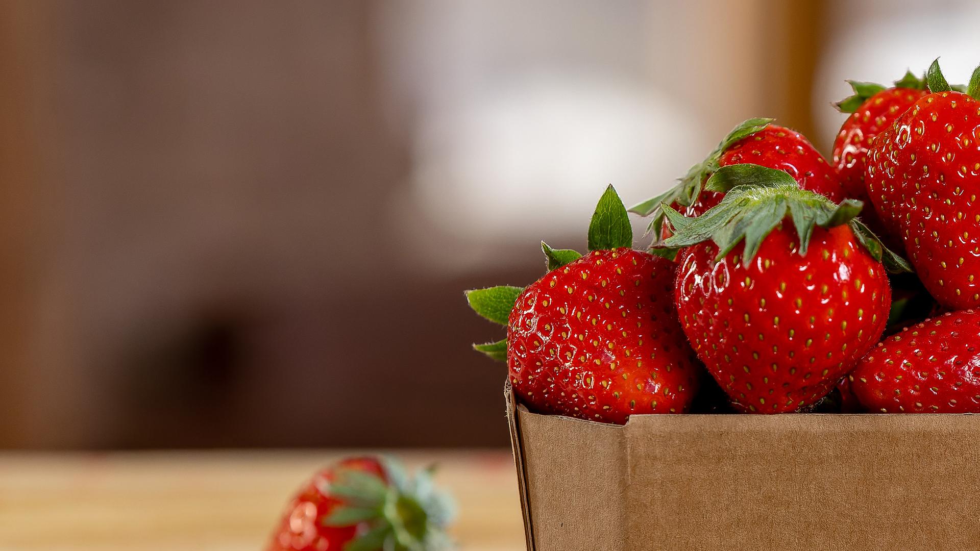 Im Anschnitt rechts eine braune Pappschale voll mit frischen reifen Erdbeeren.