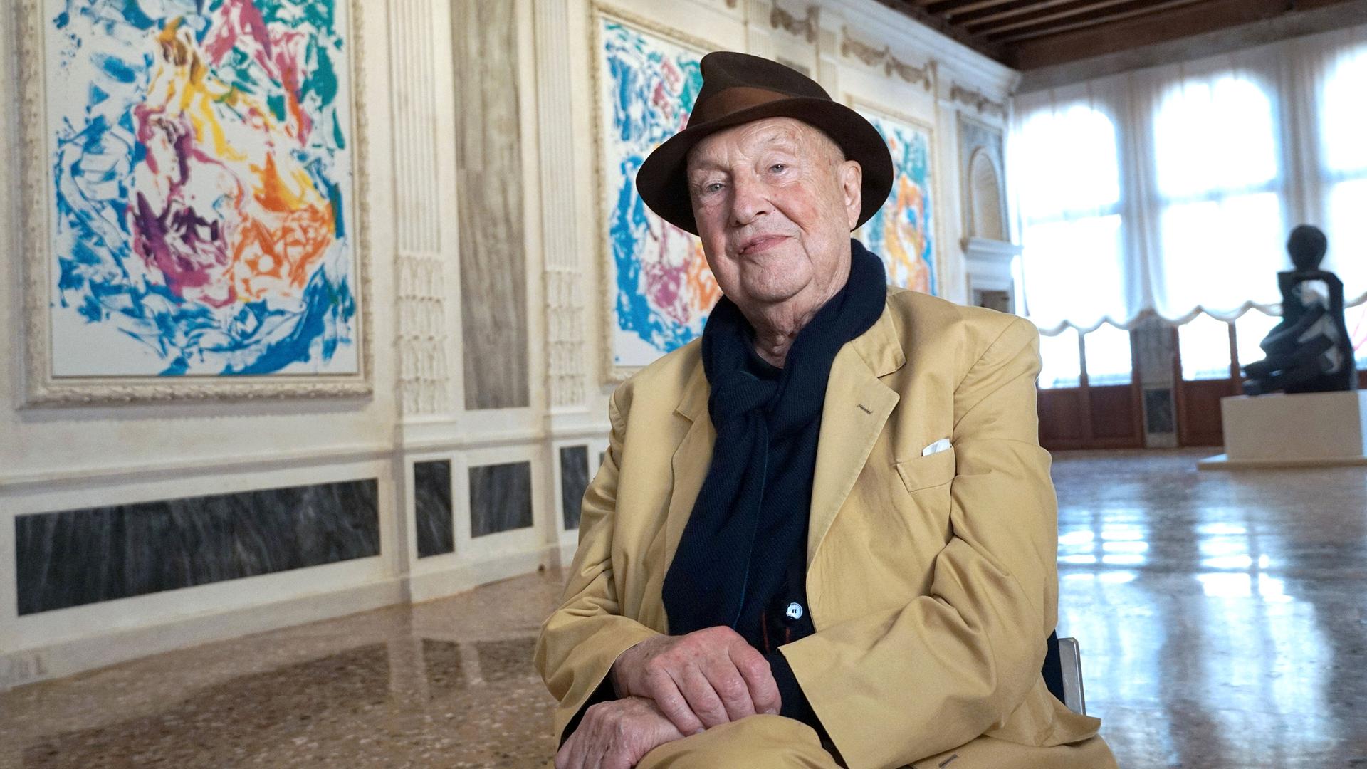 Der Maler Georg Baselitz sitzt auf einem Stuhl in einer Ausstellung mit seinen Bildern