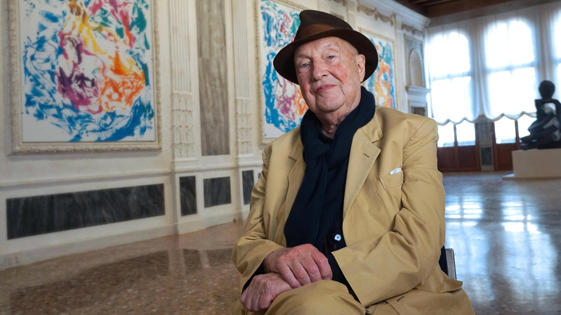 Der Maler Georg Baselitz sitzt auf einem Stuhl in einer Ausstellung mit seinen Bildern