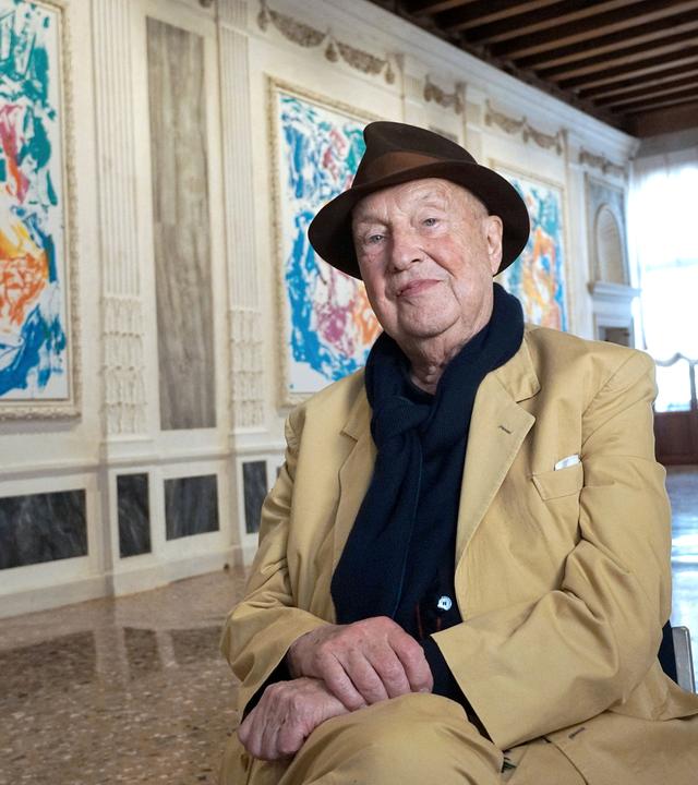 Der Maler Georg Baselitz sitzt auf einem Stuhl in einer Ausstellung mit seinen Bildern