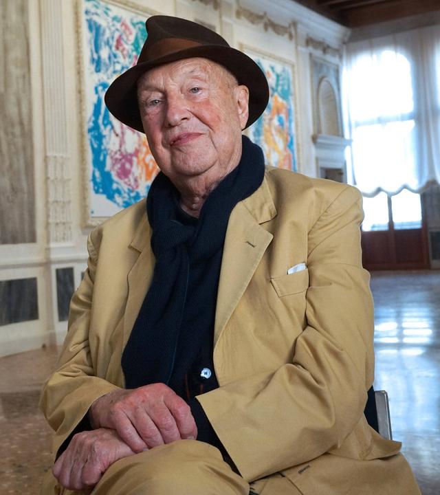 Der Maler Georg Baselitz sitzt auf einem Stuhl in einer Ausstellung mit seinen Bildern