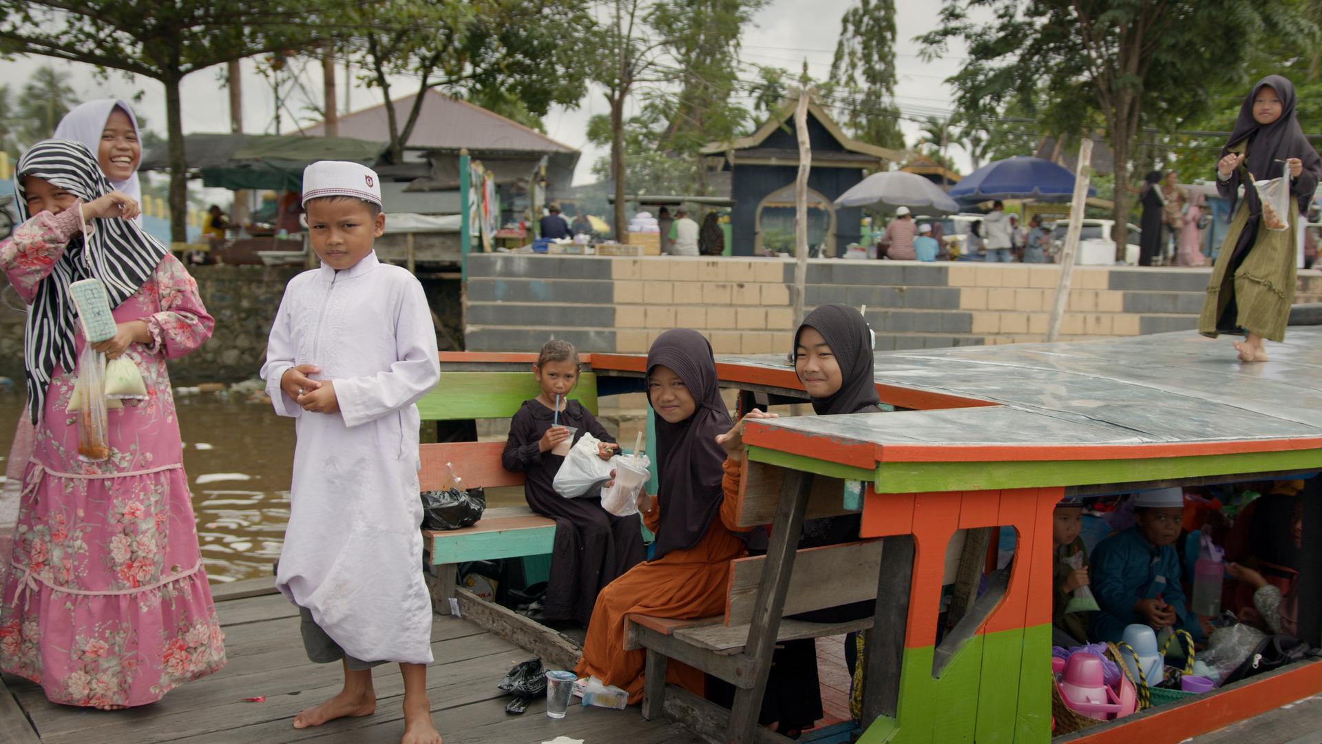 Mehrere Kinder auf einer Barkasse, Mädchen in Kleid und meist mit Kopftuch, Jungen in langem Gewand und mit indonesischer Kopfbedeckung. Im Hintergrund eine Treppe, die zum Wasser führt, sowie mehrere Gebäude, Verkaufsstände und Passanten