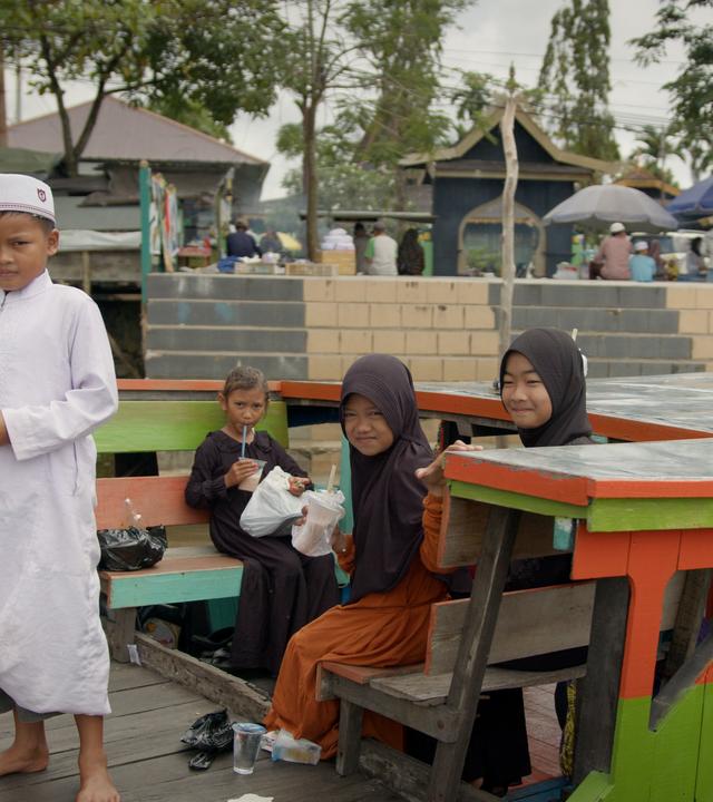 Mehrere Kinder auf einer Barkasse, Mädchen in Kleid und meist mit Kopftuch, Jungen in langem Gewand und mit indonesischer Kopfbedeckung. Im Hintergrund eine Treppe, die zum Wasser führt, sowie mehrere Gebäude, Verkaufsstände und Passanten