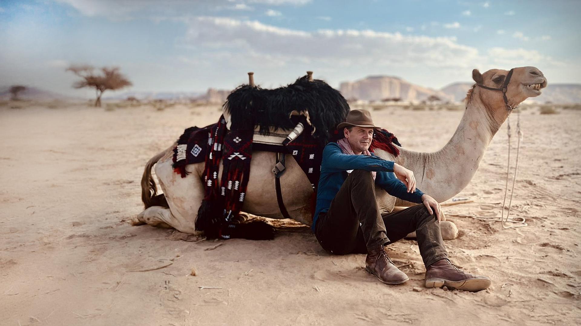 Ein Mann sitzt an ein Kamel gelehnt in einer Wüstenlandschaft.