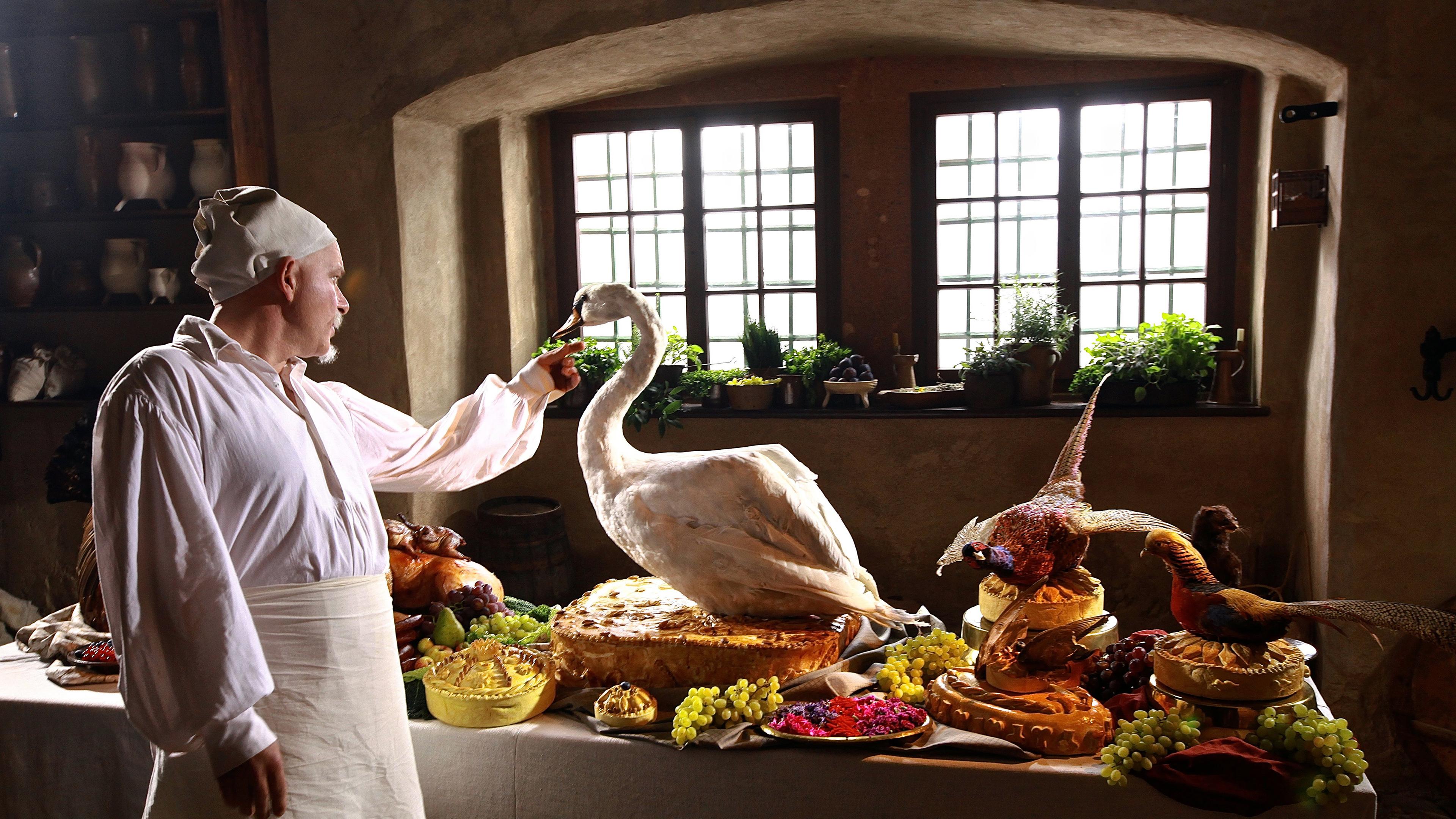 Ein mittelalterlich gewandeter Koch steht neben einem reich gedecktem Tisch mit besonders dekorativen "Schaugerichten".
