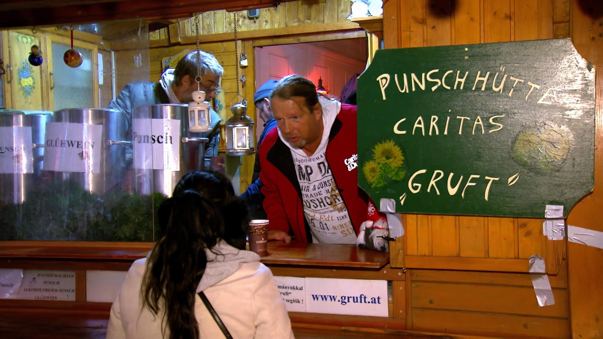 Das Bild zeigt einen Punschstand in einer Holzbaracke, die festlich dekoriert ist. Im Vordergrund ist eine Person mit langen, dunklen Haaren zu sehen, die einen weißen Kapuzenpullover trägt. Diese Person steht an einem Holzstandtisch, vor dem eine Tasse Punsch in einer Pappbecher steht.  Hinter dem Stand sind mehrere mit Beschriftungen versehene silberne Behälter zu erkennen, die entweder Punsch oder Glühwein enthalten. An einer Wand des Standes hängt ein grünes Holzschild mit der Aufschrift „Punschohütte Caritas Gruft“.   Die Atmosphäre wirkt gemütlich und einladend, mit Lichtern und weihnachtlicher Dekoration. Im Hintergrund sind einige weitere Personen zu sehen, die vermutlich ebenfalls am Stand beschäftigt sind oder darauf warten, bedient zu werden. Die gesamte Szenerie vermittelt den Eindruck eines sozialen Ereignisses, das in der Vorweihnachtszeit stattfindet.