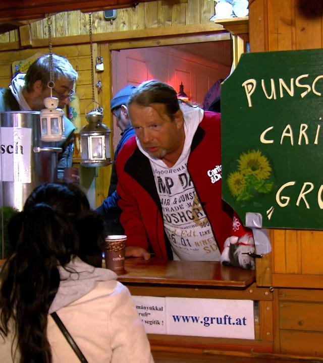 Das Bild zeigt einen Punschstand in einer Holzbaracke, die festlich dekoriert ist. Im Vordergrund ist eine Person mit langen, dunklen Haaren zu sehen, die einen weißen Kapuzenpullover trägt. Diese Person steht an einem Holzstandtisch, vor dem eine Tasse Punsch in einer Pappbecher steht.  Hinter dem Stand sind mehrere mit Beschriftungen versehene silberne Behälter zu erkennen, die entweder Punsch oder Glühwein enthalten. An einer Wand des Standes hängt ein grünes Holzschild mit der Aufschrift „Punschohütte Caritas Gruft“.   Die Atmosphäre wirkt gemütlich und einladend, mit Lichtern und weihnachtlicher Dekoration. Im Hintergrund sind einige weitere Personen zu sehen, die vermutlich ebenfalls am Stand beschäftigt sind oder darauf warten, bedient zu werden. Die gesamte Szenerie vermittelt den Eindruck eines sozialen Ereignisses, das in der Vorweihnachtszeit stattfindet.