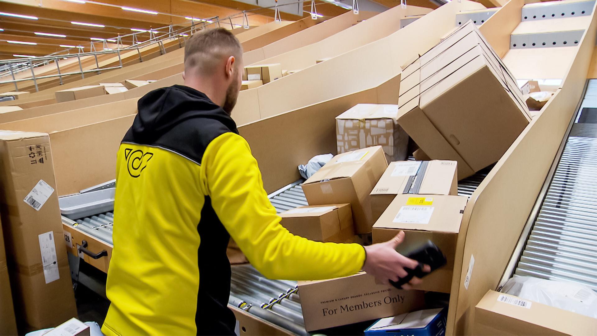 Das Bild zeigt einen Mitarbeiter in einem Paketverteilzentrum. Er trägt eine schwarz-gelbe Arbeitsjacke und ist dabei, Kartons zu sortieren. Die Umgebung ist geprägt von lange gestreckten Förderbändern, auf denen zahlreiche Päckchen und Pakete liegen. Die Pakete sind in verschiedenen Größen und Formen, einige sind beschriftet, während andere versiegelt sind. Im Hintergrund sind weitere Reihen von Paketen sichtbar. Die Decke des Zentrums ist aus Holz und es gibt eine helle Beleuchtung. Der Mitarbeiter ist von der Seite zu sehen und scheint in konzentrierter Arbeit zu sein, während er ein Paket in der Hand hält.