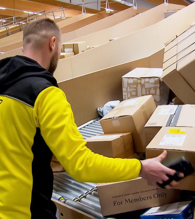 Das Bild zeigt einen Mitarbeiter in einem Paketverteilzentrum. Er trägt eine schwarz-gelbe Arbeitsjacke und ist dabei, Kartons zu sortieren. Die Umgebung ist geprägt von lange gestreckten Förderbändern, auf denen zahlreiche Päckchen und Pakete liegen. Die Pakete sind in verschiedenen Größen und Formen, einige sind beschriftet, während andere versiegelt sind. Im Hintergrund sind weitere Reihen von Paketen sichtbar. Die Decke des Zentrums ist aus Holz und es gibt eine helle Beleuchtung. Der Mitarbeiter ist von der Seite zu sehen und scheint in konzentrierter Arbeit zu sein, während er ein Paket in der Hand hält.