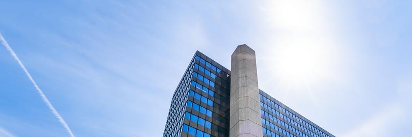 Aus niedriger Perspektive blickt man auf das ZDF-Hochhaus mit seiner charakteristischen Fensterstruktur und einem zentralen Betonelement. Im Vordergrund rahmen Bäume das Gebäude ein und verstärken die monumentale Wirkung.