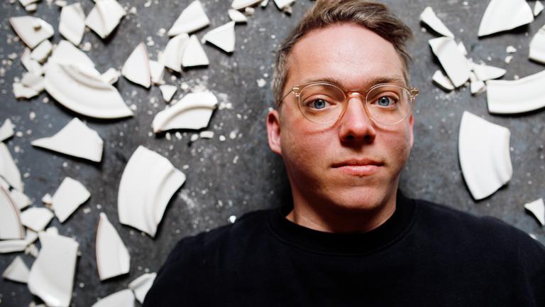 Janik liegt auf dem Boden und schaut in die Kamera. Um ihn herum sind Scherben zu sehen.