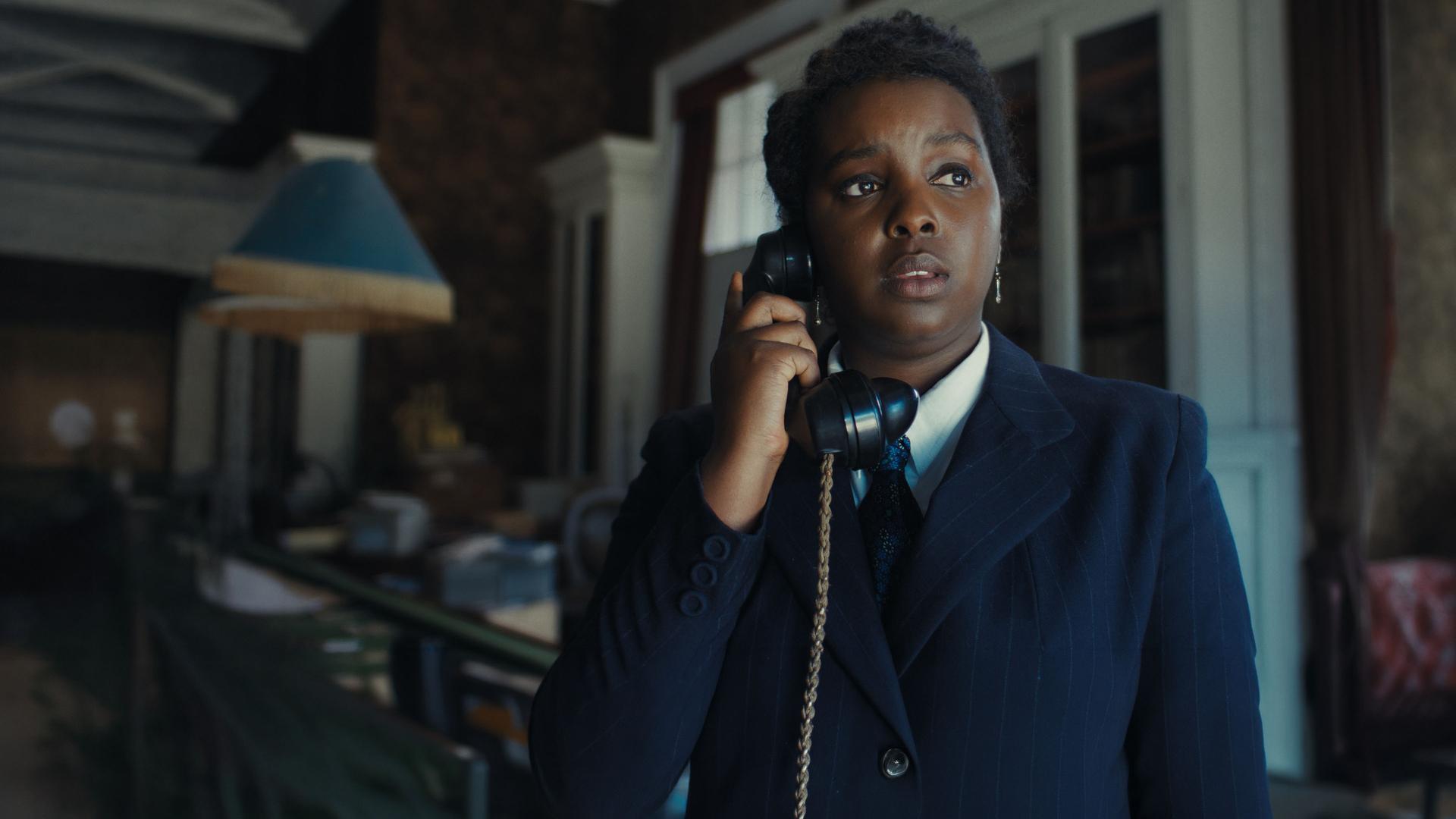 "This Is Not a Murder Mystery - Geschenk": DC Quant (Donna Banya) befindet sich in einem Raum. Sie hält sich mit der rechten Hand einen Telefonhörer ans Ohr.