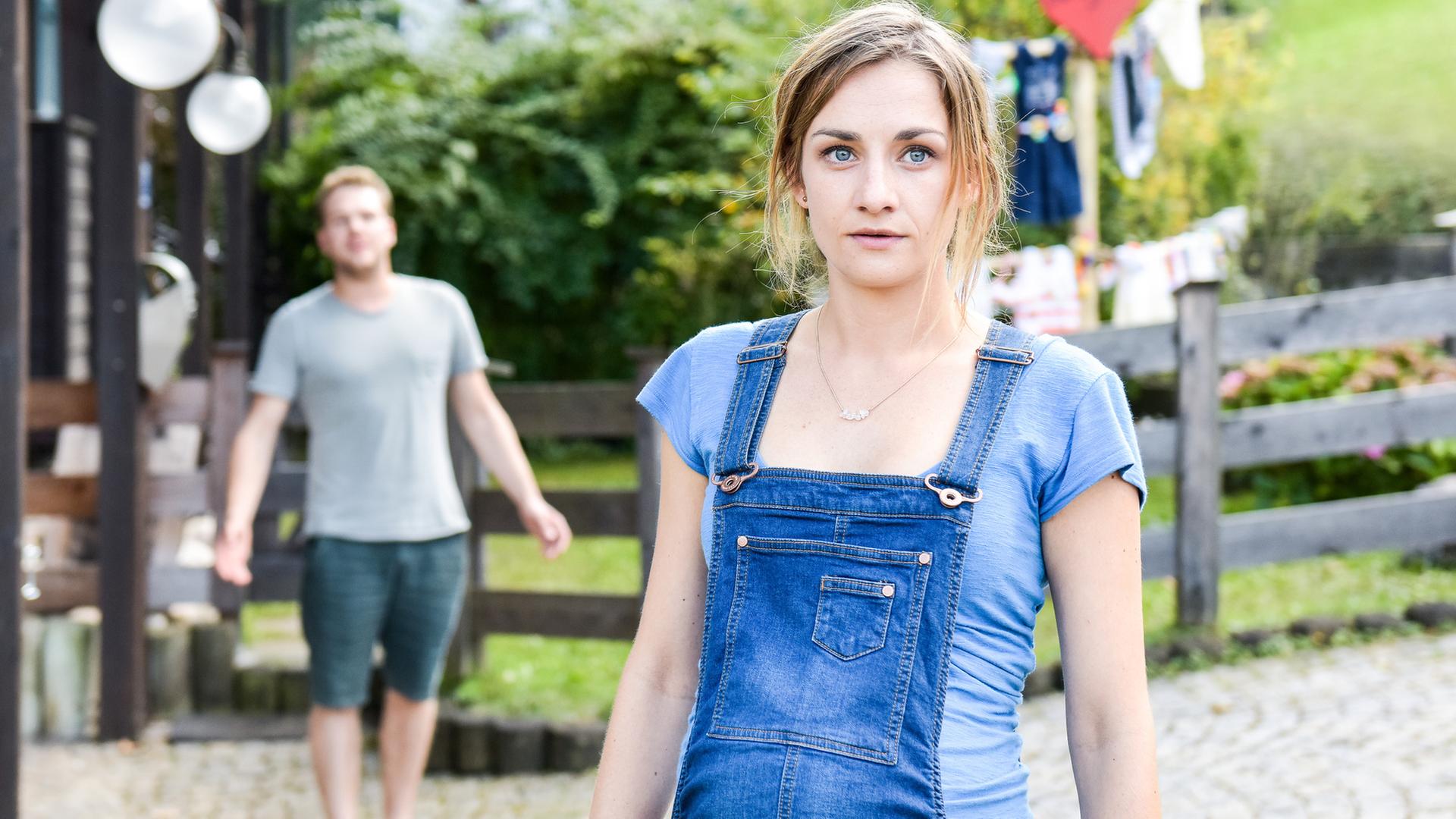 "Lena Lorenz - Geschwisterliebe": Miriam (Karoline Teska) steht vor einem Haus im Bildvordergrund. Versetzt hinter ihr in der Unschärfe steht Benjamin (Ben Münchow) und scheint ihr etwas zuzurufen.