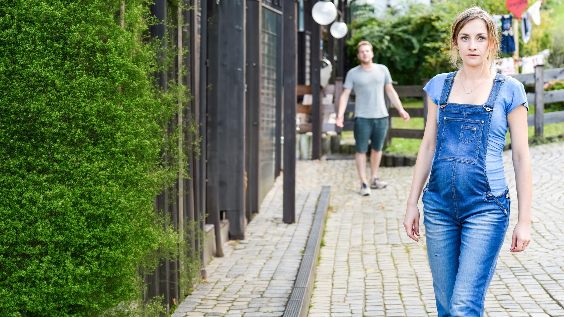 "Lena Lorenz - Geschwisterliebe": Miriam (Karoline Teska) steht vor einem Haus im Bildvordergrund. Versetzt hinter ihr in der Unschärfe steht Benjamin (Ben Münchow) und scheint ihr etwas zuzurufen.