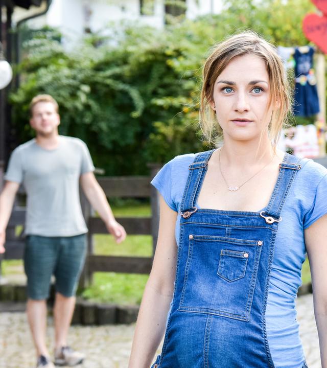 "Lena Lorenz - Geschwisterliebe": Miriam (Karoline Teska) steht vor einem Haus im Bildvordergrund. Versetzt hinter ihr in der Unschärfe steht Benjamin (Ben Münchow) und scheint ihr etwas zuzurufen.