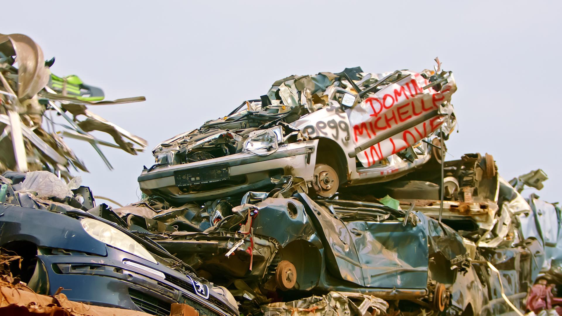 Das Bild zeigt einen Schrottplatz, auf dem zahlreiche alte, beschädigte Autos übereinander gestapelt sind. Es sind verschiedene Fahrzeugmodelle sichtbar, die stark verrostet und verformt sind. Ein Auto in der Mitte trägt rote Schrift mit dem Text "Dominique Michelle in Love." Der Hintergrund ist meist wolkenverhangen und wirkt trüb. Der Anblick ist geprägt von einer chaotischen Ansammlung von Metall und teilweise auch anderen Materialien, die auf eine mangelhafte Abfallwirtschaft hinweisen. Der Kontext des Bildes behandelt die Problematik der Müllentsorgung und das Thema einer "Gesellschaft ohne Müll."