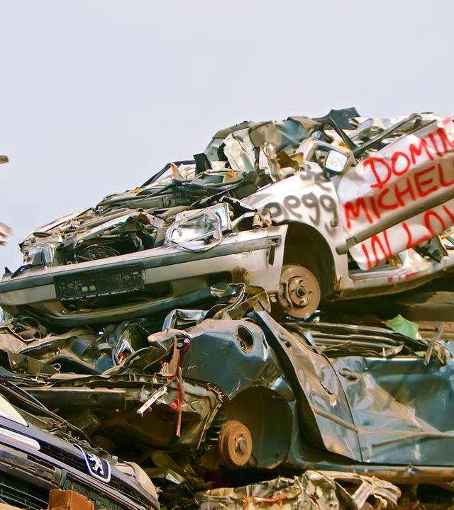 Das Bild zeigt einen Schrottplatz, auf dem zahlreiche alte, beschädigte Autos übereinander gestapelt sind. Es sind verschiedene Fahrzeugmodelle sichtbar, die stark verrostet und verformt sind. Ein Auto in der Mitte trägt rote Schrift mit dem Text "Dominique Michelle in Love." Der Hintergrund ist meist wolkenverhangen und wirkt trüb. Der Anblick ist geprägt von einer chaotischen Ansammlung von Metall und teilweise auch anderen Materialien, die auf eine mangelhafte Abfallwirtschaft hinweisen. Der Kontext des Bildes behandelt die Problematik der Müllentsorgung und das Thema einer "Gesellschaft ohne Müll."