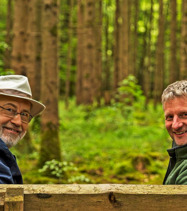 Harald  Lesch und Joachim Keßler sitzen auf einer Bank im Wald
