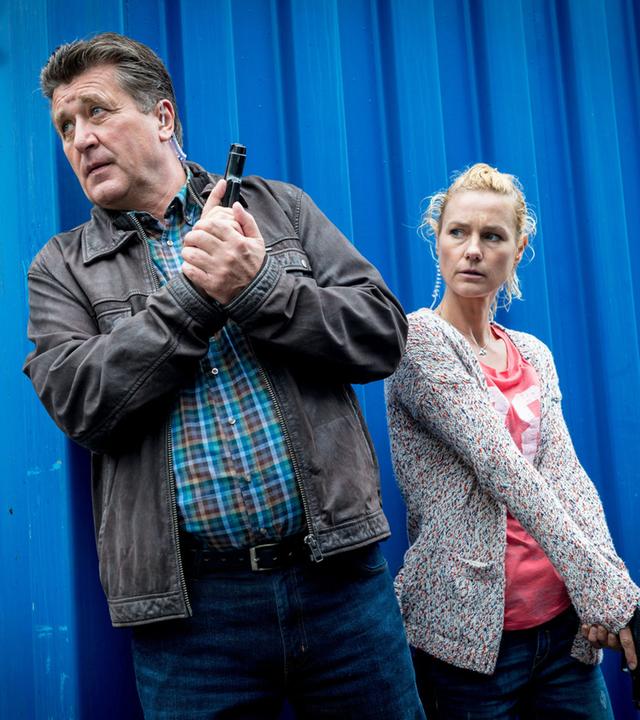 "Notruf Hafenkante - Gestohlenes Leben": Hans Moor (Bruno F. Apitz) und Franzi Jung (Rhea Harder-Vennewald) stehen in Deckung hinter einem blauen Container. Beide halten ihre Waffen einsatzbereit. Sie sind in Zivil.
