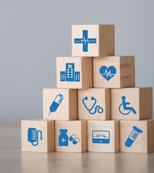 Auf einem Tisch sind Holzklötze zu einer kleinen Pyramide aufgebaut. Die Klötze tragen blaue medizinische Symbole wie EKG, Spritze, Tabletten, Rollstuhl und Klinikgebäude. Das Bild visualisiert die vielfältigen Elemente des Gesundheitswesens und deren Zusammenspiel.