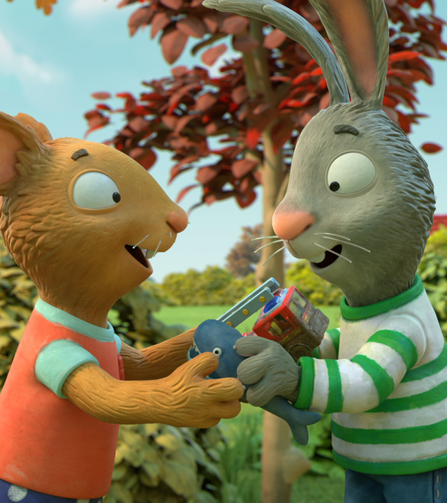 Draußen im Grünen: Die Maus Posy und ihr Freund Pip, das Kaninchen, stehen sich gegenüber. Sie halten ihre Hände aneinander. In Posys linker Hand befindet sich ein Feuerwehrauto und in Pips rechter Hand ein kleiner Hai. Sie lächeln beide erwartungsvoll und freudig. Im Hintergrund sind Sträucher zu sehen.