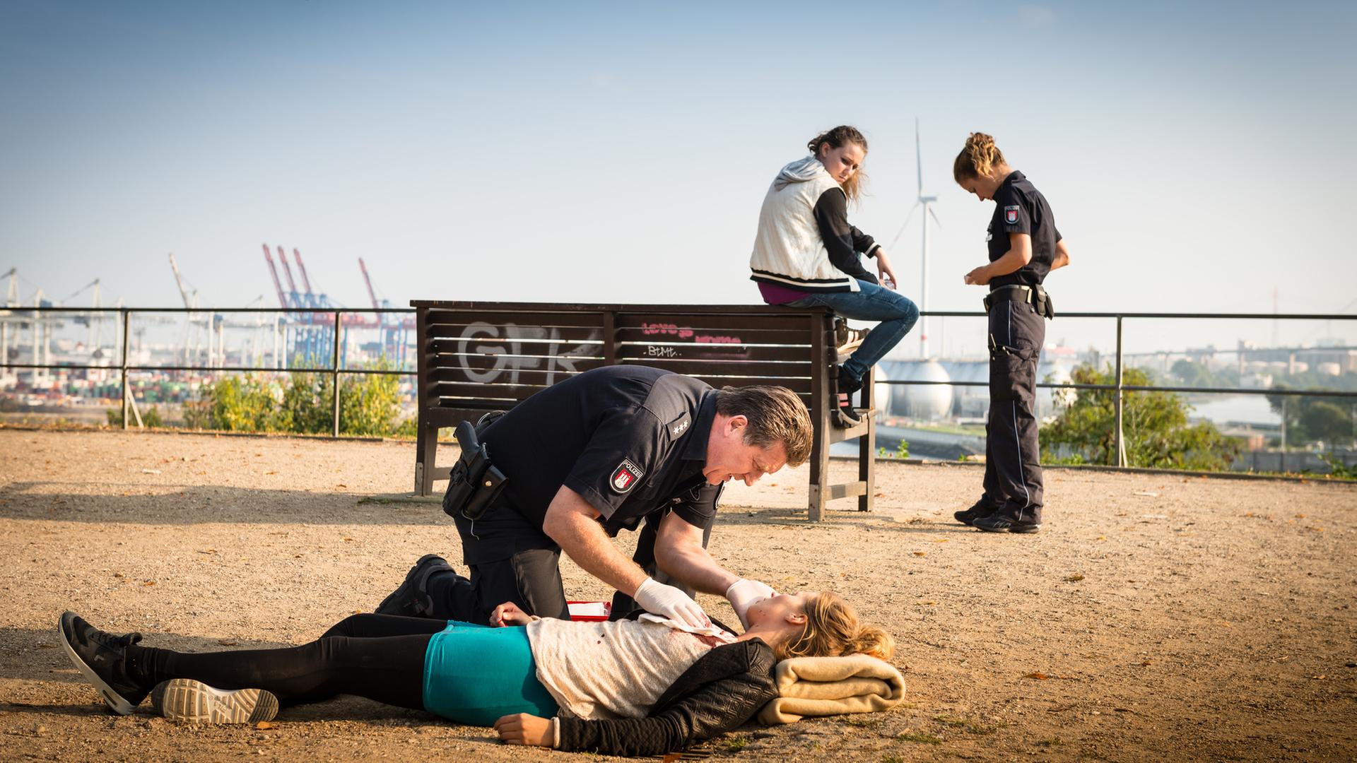 "Notruf Hafenkante - Getrieben": Der Polizeibeamte Hans Moor (Bruno F. Apitz) kniet über Annalena Stapel (Zarina Zöller), die auf dem Boden liegt. Die Polizeibeamtin Franzi Jung (Rhea Harder-Vennewald) steht bei Britta Hillemann (Vivien Fröhlich.).