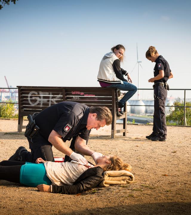 "Notruf Hafenkante - Getrieben": Der Polizeibeamte Hans Moor (Bruno F. Apitz) kniet über Annalena Stapel (Zarina Zöller), die auf dem Boden liegt. Die Polizeibeamtin Franzi Jung (Rhea Harder-Vennewald) steht bei Britta Hillemann (Vivien Fröhlich.).