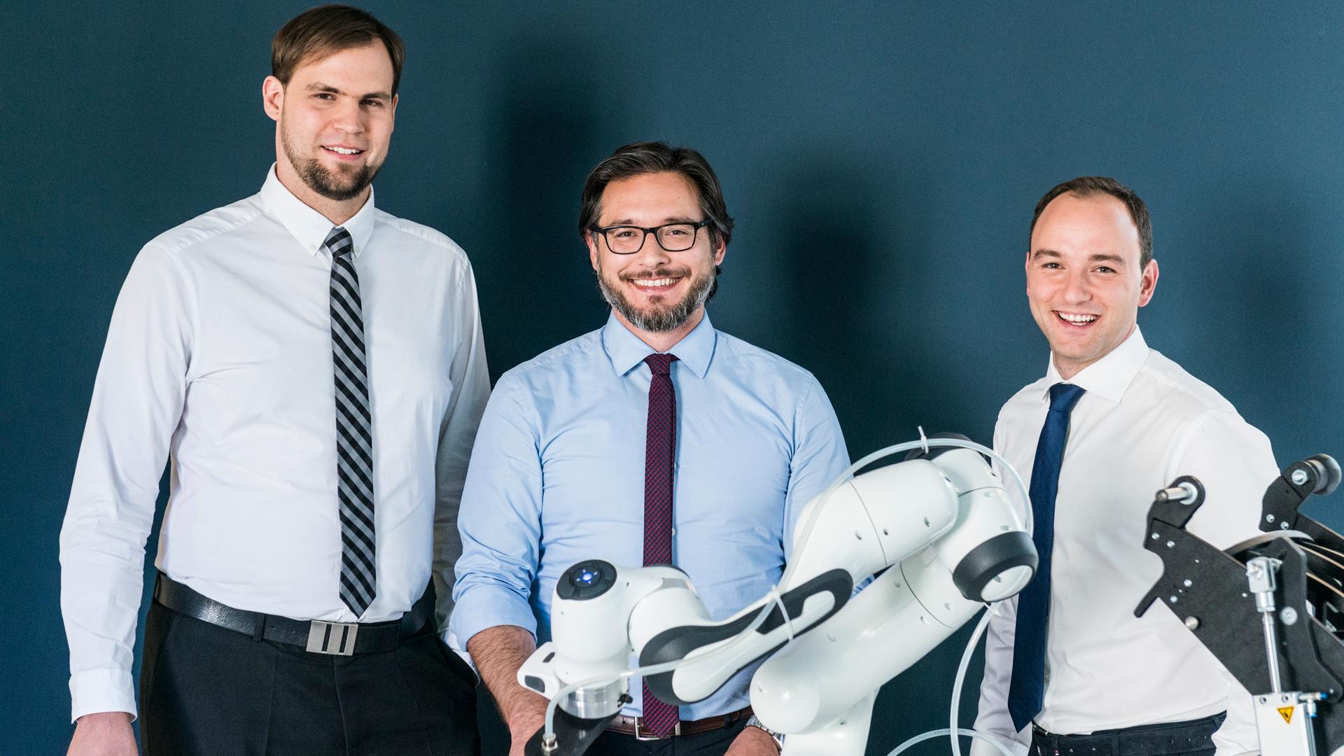 Drei Männer stehen hinter einem Roboterarm und lachen in die Kamera.